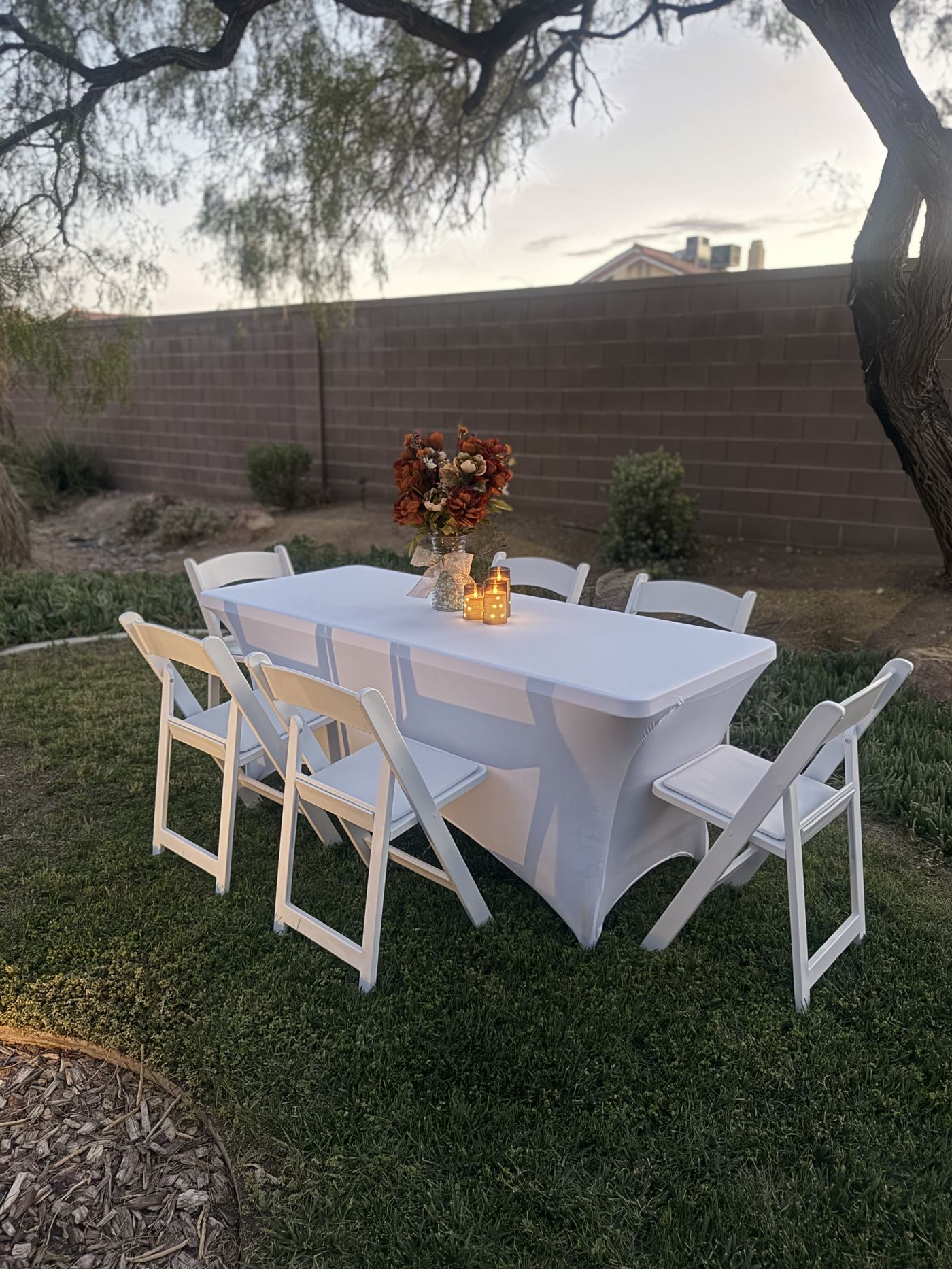 Six foot rectangle table and white folding chairs set on a lawn at dusk